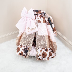 May include: A pink and brown car seat cover with a large pink bow and the name "Davie" embroidered on it. The cover features a cow print, pink roses, and gold glitter fabric.