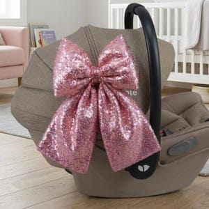 May include: A large, pink sequined bow attached to a tan baby car seat. The bow is the focal point, with its shimmering surface catching the light. The car seat has a black handle and is set in a room with a crib and other baby items.