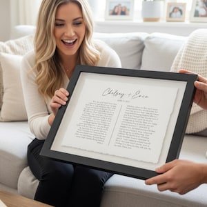 May include: A framed print featuring the names "Chelsey + Eric" and a handwritten letter. The black frame surrounds the white paper with the text. The print is being held by two people.