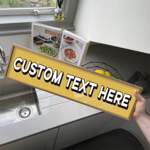 May include: A yellow sign with black text that reads "CUSTOM TEXT HERE". The sign is hanging on a kitchen counter.