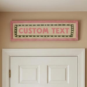 May include: A rectangular sign with the words "CUSTOM TEXT" in pink letters. The sign has a pink frame with a beaded edge and a green and black border. The sign is mounted above a white door.