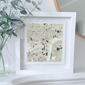 May include: A framed black and white map print of London. The map, set within a white frame, shows the city's layout. The print is displayed on a white surface, with a vase of greenery in the background, adding a touch of nature.
