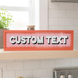 May include: A rectangular sign with the words "CUSTOM TEXT" in bold white letters with black drop shadows. The sign has a pink background with a scalloped edge and is framed in a coral-coloured border. The sign is displayed on a wooden shelf.