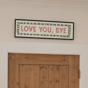 May include: A framed sign with the words "LOVE YOU, BYE" in pink. The sign has a white background and a green border. It is mounted above a wooden door with a natural finish.