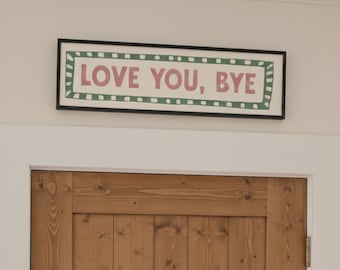 May include: A rectangular framed sign with the text "LOVE YOU, BYE" in pink letters. The sign has a white background with a green border and is mounted above a wooden door.
