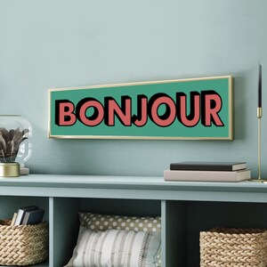 Puede incluir: Un letrero rectangular verde azulado con marco dorado. El letrero dice "BONJOUR" en letras rojas grandes y en negrita.
