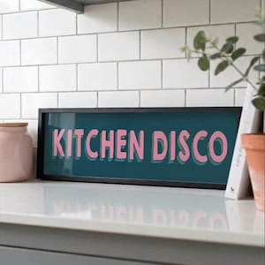 May include: A framed sign displaying the words "KITCHEN DISCO" in pink, set against a teal background. The sign is in a black frame, resting on a white surface. A pink jar with a wooden lid is on the left, and a potted plant is on the right.