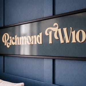 May include: A rectangular sign reading "Richmond TW10" in a cream-coloured, stylised font. The sign is set against a dark teal background and framed in black, mounted on a blue wall.