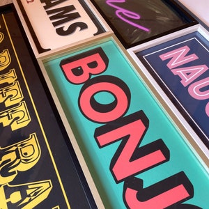 Puede incluir: Primer plano de una pared con carteles enmarcados. Los carteles muestran las palabras "BONJOUR", "NAUGHTY" y "FIRST RAY" en letras negritas y coloridas.
