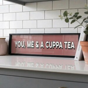 May include: A rectangular framed sign with the words "YOU, ME & A CUPPA TEA" in white, outlined in black, on a red background. The black frame sits on a white surface, with a white tiled wall in the background.