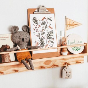 May include: A wooden shelf with a gray stuffed mouse, a book titled "I Want My Hat Back", a clipboard with a botanical print, a wooden bunny toy, a round wooden sign with the name "Hudson" and a wooden tag with the letter "A" and an alpaca illustration.