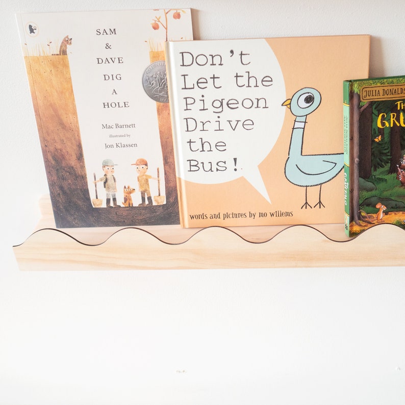 Peut inclure: Trois livres pour enfants sur une &eacute;tag&egrave;re en bois. Le livre du milieu est intitul&eacute; "Ne laissez pas le pigeon conduire le bus !" et pr&eacute;sente un pigeon bleu avec une bulle de dialogue. Le livre de gauche est intitul&eacute; "Sam & Dave creusent un trou" et pr&eacute;sente deux enfants en train de creuser un trou. Le livre de droite est intitul&eacute; "La fille qui a bu la lune" et pr&eacute;sente une fille dans une for&ecirc;t.