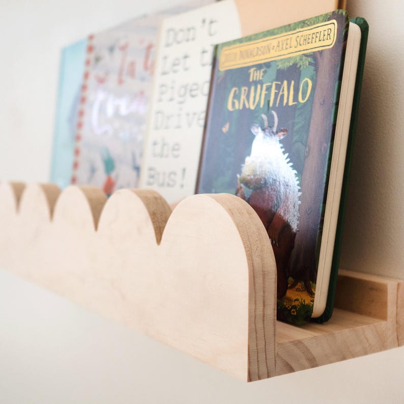 May include: A wooden cloud-shaped bookshelf holds several children's books. The books have colorful covers, including one titled "The Gruffalo." The shelf is made of light-colored wood and is mounted on a white wall.