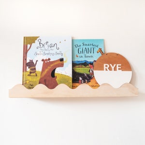 Peut inclure: Deux livres pour enfants, "Brian: The Smelly Bear Smells Something Smelly" et "The Smartest Giant in Town", sont expos&eacute;s sur une &eacute;tag&egrave;re en bois avec une pancarte ronde en bois blanc et brun qui dit "RYE" en lettres blanches.