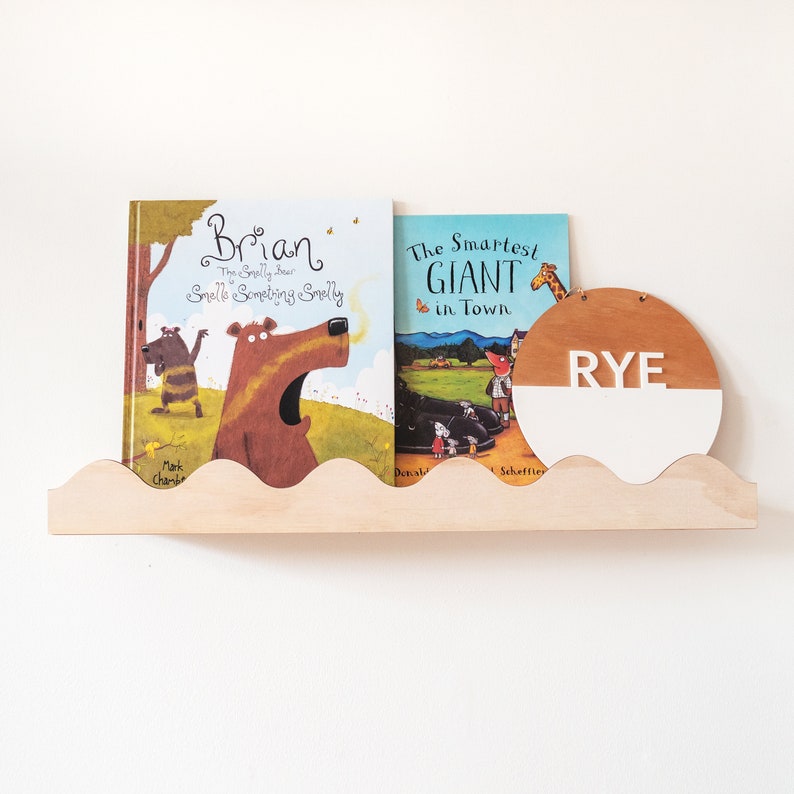 Peut inclure: Deux livres pour enfants, "Brian: The Smelly Bear Smells Something Smelly" et "The Smartest Giant in Town", sont expos&eacute;s sur une &eacute;tag&egrave;re en bois avec une pancarte ronde en bois blanc et brun qui dit "RYE" en lettres blanches.