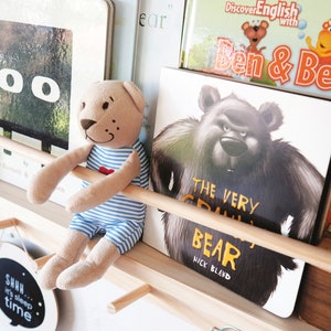 May include: A wooden shelf with a variety of children's books, including "The Very Cranky Bear" by Nick Bland, "Discover English with Ben & Bella", and a book with a black square with two white circles. A brown teddy bear wearing a blue and white striped shirt is sitting on the shelf.