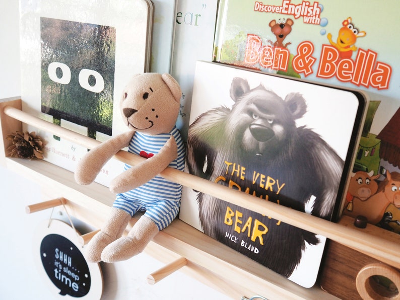 May include: A wooden shelf with a variety of children's books, including "The Very Cranky Bear" by Nick Bland, "Discover English with Ben & Bella", and a book with a black square with two white circles. A brown teddy bear wearing a blue and white striped shirt is sitting on the shelf.