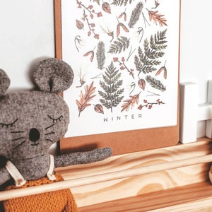 May include: A gray felt mouse toy with closed eyes and a stitched-on nose sits on a wooden shelf. The shelf also features a wooden bunny toy, a white wooden letter, and a framed print with a winter-themed design and the word "WINTER" at the bottom.