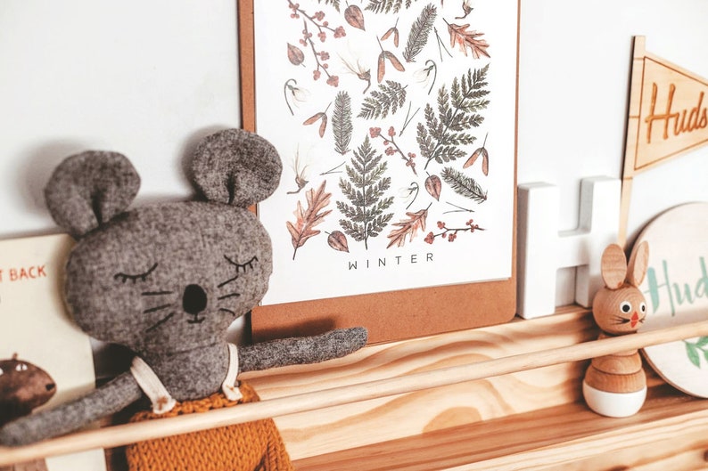 May include: A gray felt mouse toy with closed eyes and a stitched-on nose sits on a wooden shelf. The shelf also features a wooden bunny toy, a white wooden letter, and a framed print with a winter-themed design and the word "WINTER" at the bottom.