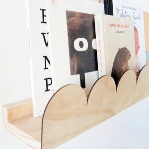 May include: A wooden cloud-shaped bookshelf holds several children's books. The shelf is made of light-colored wood and has a scalloped edge. The books have colorful covers and titles, including "I Want My Hat Back."