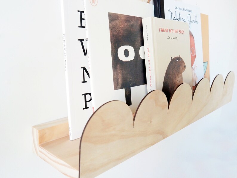 May include: A wooden cloud-shaped bookshelf holds several children's books. The shelf is made of light-colored wood and has a scalloped edge. The books have colorful covers and titles, including "I Want My Hat Back."