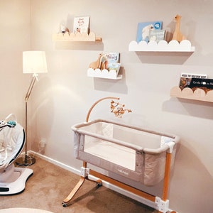 May include: A nursery scene featuring a light-colored crib with a mobile, cloud-shaped shelves holding books and wooden animal figurines, and a floor lamp. The crib has a beige fabric and wooden frame. The shelves are white and natural wood.