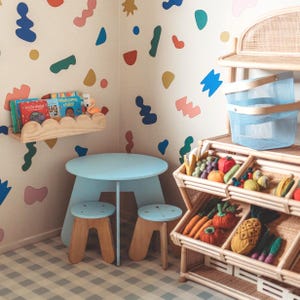 May include: A light blue children's table and two stools with wooden legs. A wooden shelf holds books and a flamingo figurine. A multi-colored wall design and a rattan display with toy fruits and vegetables complete the scene.