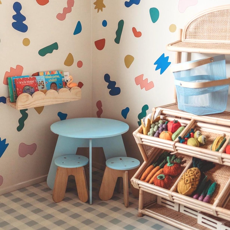 May include: A light blue children's table and two stools with wooden legs. A wooden shelf holds books and a flamingo figurine. A multi-colored wall design and a rattan display with toy fruits and vegetables complete the scene.