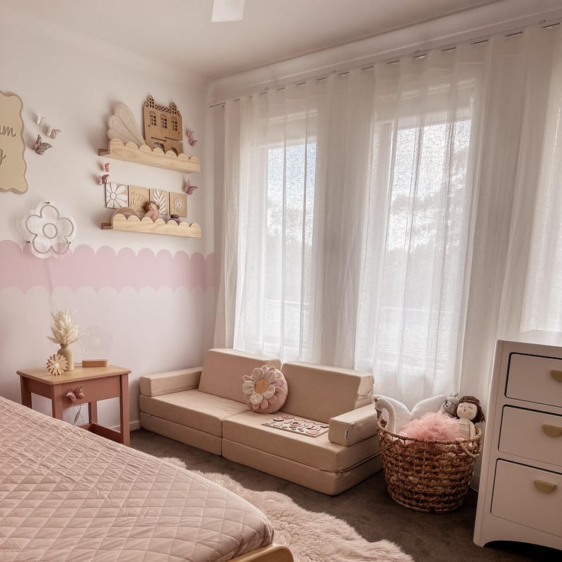 May include: A light beige modular sofa with a daisy-shaped pillow sits on a light-colored rug. A small wooden table with a pink finish is next to a bed with a pink quilted comforter. The room has white curtains and a white dresser.