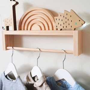 May include: A wooden wall-mounted shelf with a hanging rod, featuring a rainbow-shaped wooden toy, a wooden giraffe toy, and a wooden hedgehog toy. Three white hangers are attached to the rod.