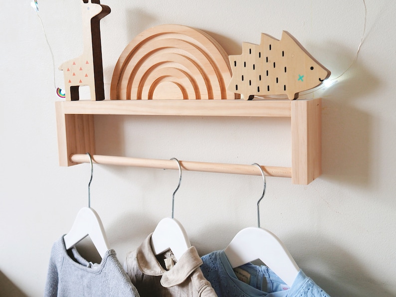 May include: A wooden wall-mounted shelf with a hanging rod, featuring a rainbow-shaped wooden toy, a wooden giraffe toy, and a wooden hedgehog toy. Three white hangers are attached to the rod.