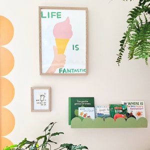 May include: A framed print with the words "LIFE IS FANTASTIC" and an ice cream cone illustration. Below, a smaller framed print and a green cloud-shaped shelf holding children's books are visible.