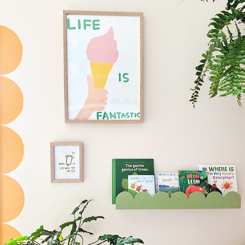 May include: A framed print with the words "LIFE IS FANTASTIC" and an ice cream cone illustration. Below, a smaller framed print and a green cloud-shaped shelf holding children's books are visible.