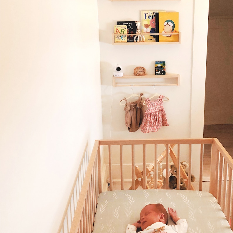 May include: A white crib with a green and white patterned sheet. A baby is sleeping in the crib. There are wooden shelves above the crib with books and a baby monitor.