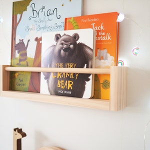 May include: A wooden shelf with three children's books on it. The books are titled "Brian", "The Very Cranky Bear", and "Jack and the Beanstalk". The shelf is mounted on a white wall with a string of rainbow fairy lights.