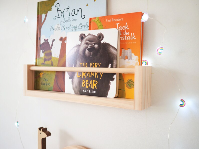 May include: A wooden shelf with three children's books on it. The books are titled "Brian", "The Very Cranky Bear", and "Jack and the Beanstalk". The shelf is mounted on a white wall with a string of rainbow fairy lights.