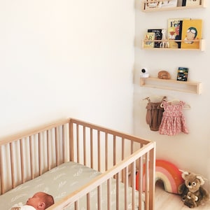 May include: A wooden crib with a white and green patterned sheet. A baby is sleeping in the crib. There are shelves on the wall with books and a baby monitor.