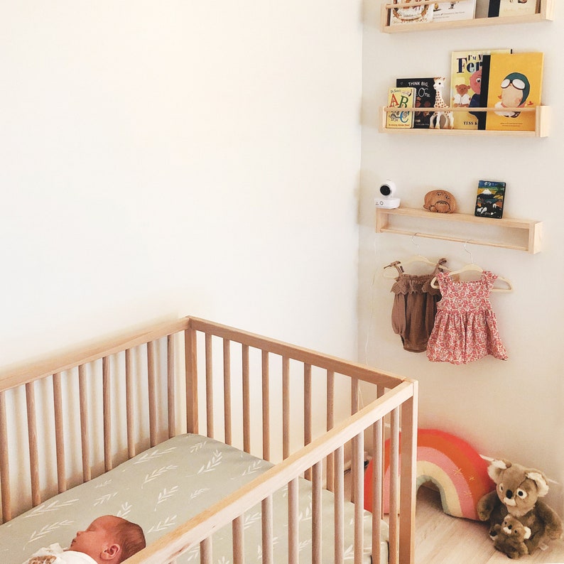 May include: A wooden crib with a white and green patterned sheet. A baby is sleeping in the crib. There are shelves on the wall with books and a baby monitor.