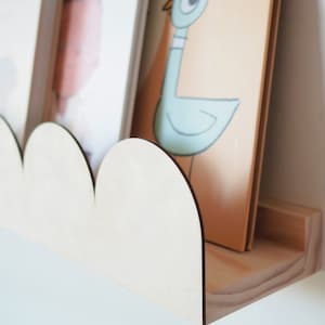 May include: A wooden cloud-shaped shelf with a natural finish, holding two framed pictures. The shelf is mounted on a white wall, showcasing a minimalist design. The shelf is made of light-colored wood.