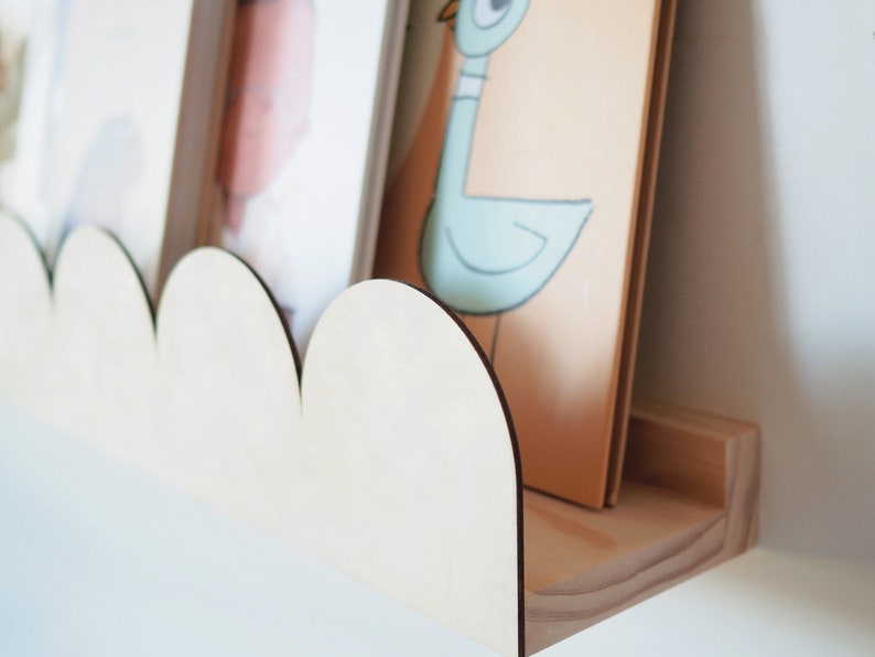 May include: A wooden cloud-shaped shelf with a natural finish, holding two framed pictures. The shelf is mounted on a white wall, showcasing a minimalist design. The shelf is made of light-colored wood.