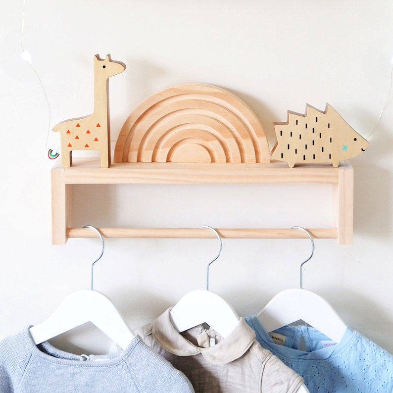 May include: Wooden shelf with a rainbow arch, a giraffe, and a hedgehog figurine. The shelf has a metal rod underneath for hanging clothes. Two white hangers with baby clothes are hanging on the rod.