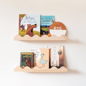 Peut inclure: Deux &eacute;tag&egrave;res en bois aux bords festonn&eacute;s, chacune contenant une collection de livres pour enfants. L'&eacute;tag&egrave;re du haut contient trois livres, dont "Brian The Broly Smell's Something Smelly" et "The Smartest Giant in Town". L'&eacute;tag&egrave;re du bas contient trois livres, dont "The Gruffalo" et "Don't Let the Pigeon Drive the Bus!". Un panneau rond en bois avec le nom "RYE" est sur l'&eacute;tag&egrave;re du haut.