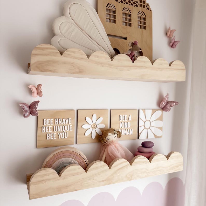 May include: Wooden cloud-shaped shelves with decorative items. Includes a wooden house, butterflies, and wooden blocks with text that reads "BEE BRAVE BEE UNIQUE BEE YOU" and "BEE A KIND HUMAN". A doll and a wooden rainbow are also present.