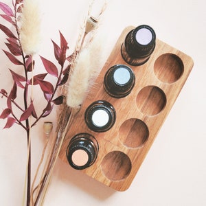May include: A wooden essential oil holder with six slots, filled with five bottles of essential oil. The bottles have different colored caps, including pink, blue, and white. The holder is on a light pink surface with dried flowers and branches.