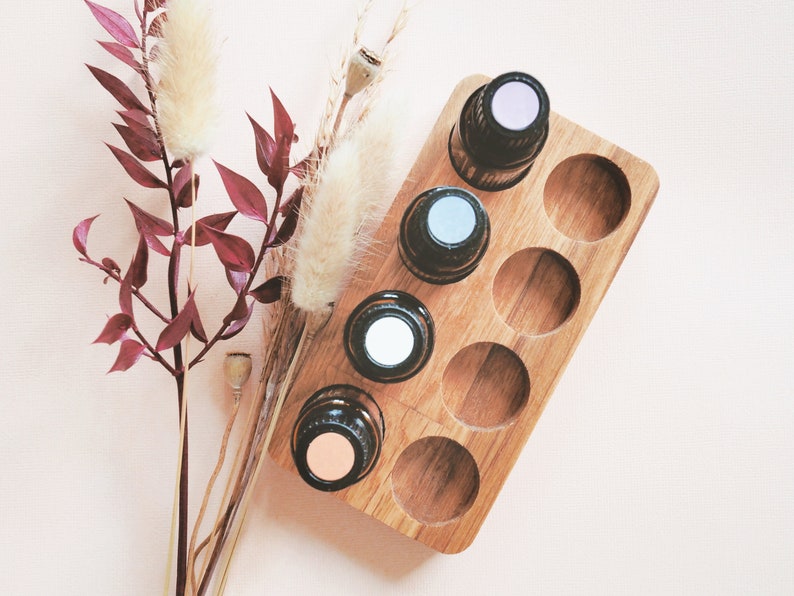 May include: A wooden essential oil holder with six slots, filled with five bottles of essential oil. The bottles have different colored caps, including pink, blue, and white. The holder is on a light pink surface with dried flowers and branches.