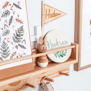 May include: A wooden shelf with a white and green round sign that says "Hudson" and a wooden flag that says "Hudson". There is a wooden toy rabbit and a framed print with a nature theme.