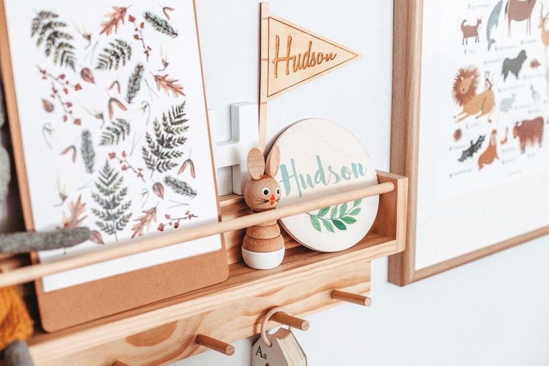 May include: A wooden shelf with a white and green round sign that says "Hudson" and a wooden flag that says "Hudson". There is a wooden toy rabbit and a framed print with a nature theme.