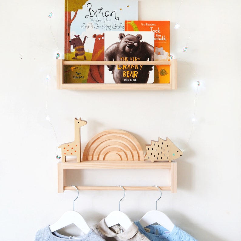 May include: A wooden shelf with a rail for hanging clothes, featuring a rainbow-shaped wooden toy, a giraffe toy, and a hedgehog toy. The shelf is decorated with a string of fairy lights and has several children's books on it, including "The Snelly Bear Smells Something Smelly" and "The Very Cranky Bear".