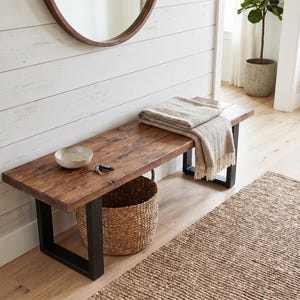 May include: A wooden bench with a dark stain and black metal legs. A beige throw blanket rests on the bench. A woven basket sits below the bench, and a small bowl and keys are on top. A woven rug is in front of the bench.