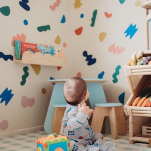 May include: A child's playroom with a light blue table and stools, a wooden shelf with books, and a multi-colored toy. The walls are decorated with colorful abstract shapes. A wooden shelving unit displays toy vegetables.